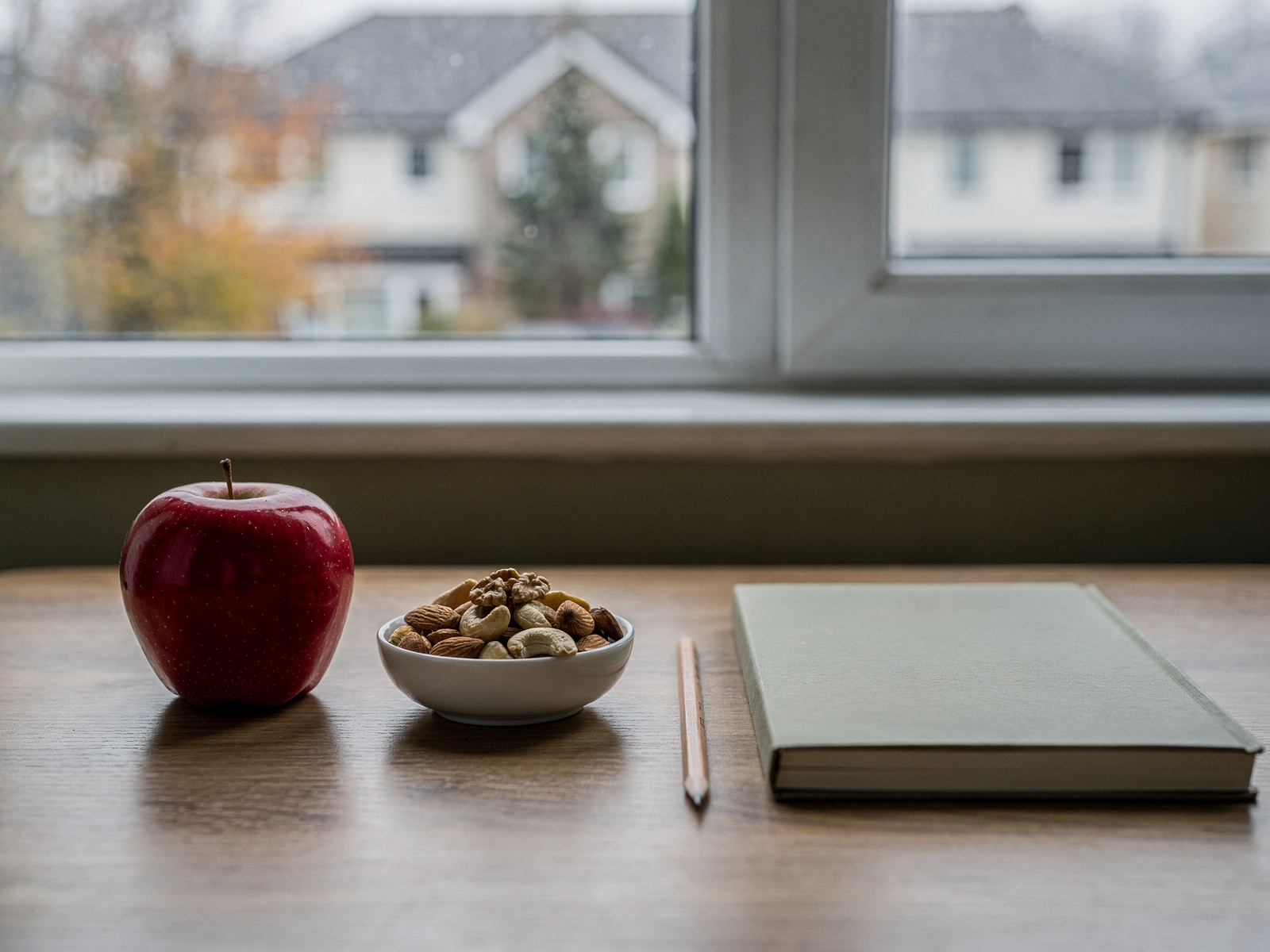 An apple and a small bowl of mixed nuts on a home desk beside a notebook, lit by a window on an overcast afternoon.
