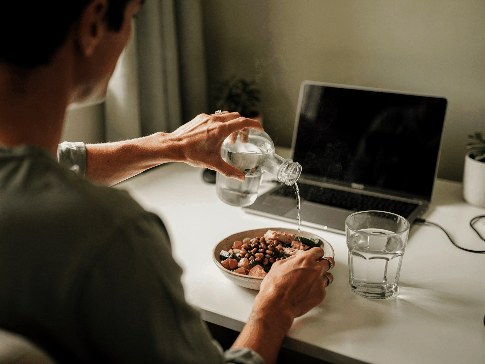 A glass of water on a tidy home desk next to a laptop and a bowl of lunch, in calm daylight.