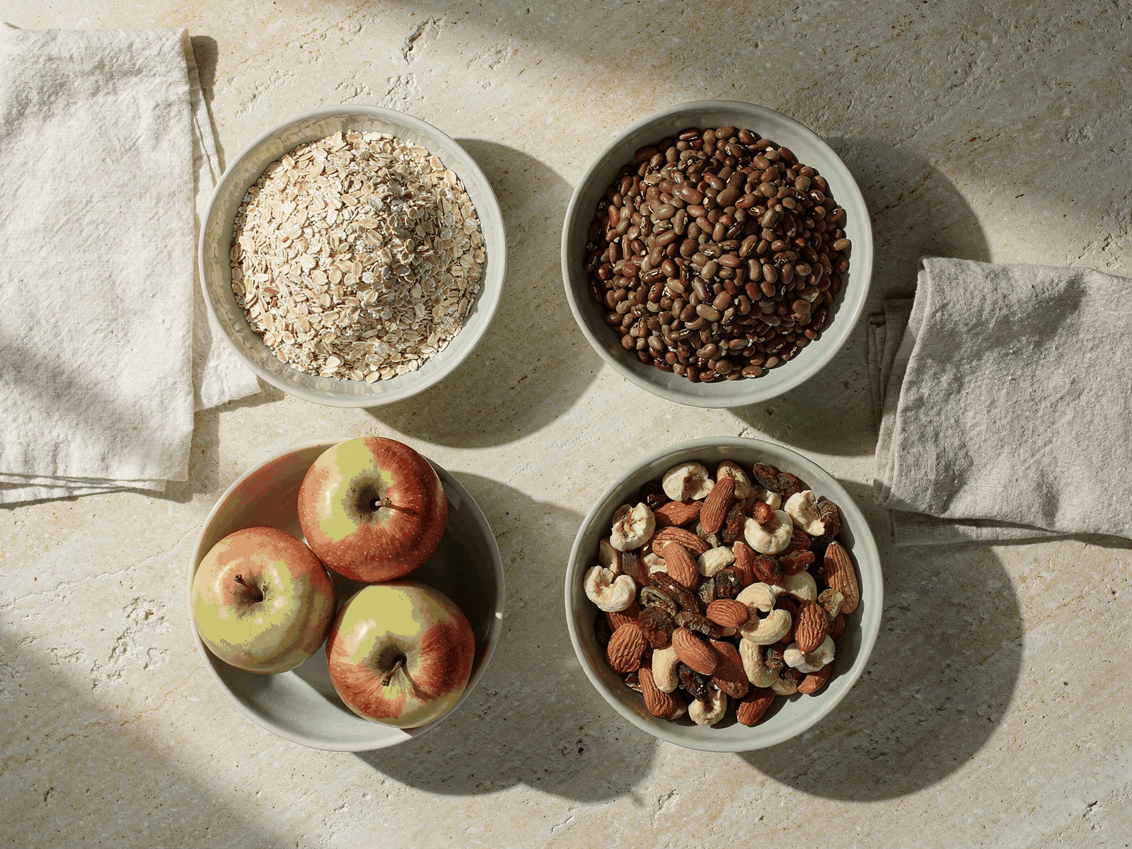 A kitchen worktop with bowls of oats, dried beans, apples and mixed nuts arranged in natural window light.