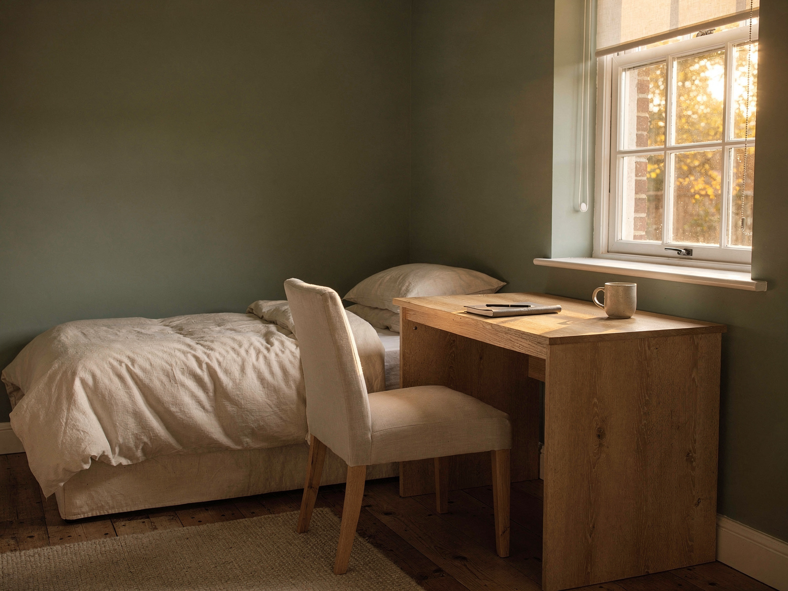 A small desk set up in the corner of a bedroom, with a window showing low late-afternoon autumn light and a made bed just behind the chair.