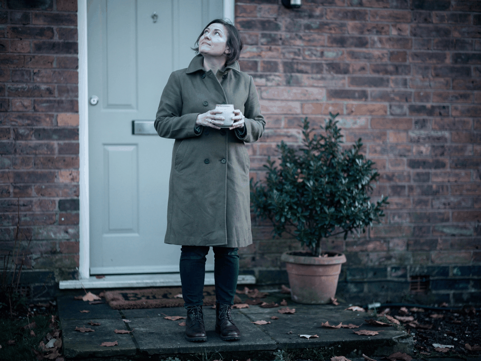A person in a coat standing on their front doorstep with a mug, in pale early-morning light before the working day starts.
