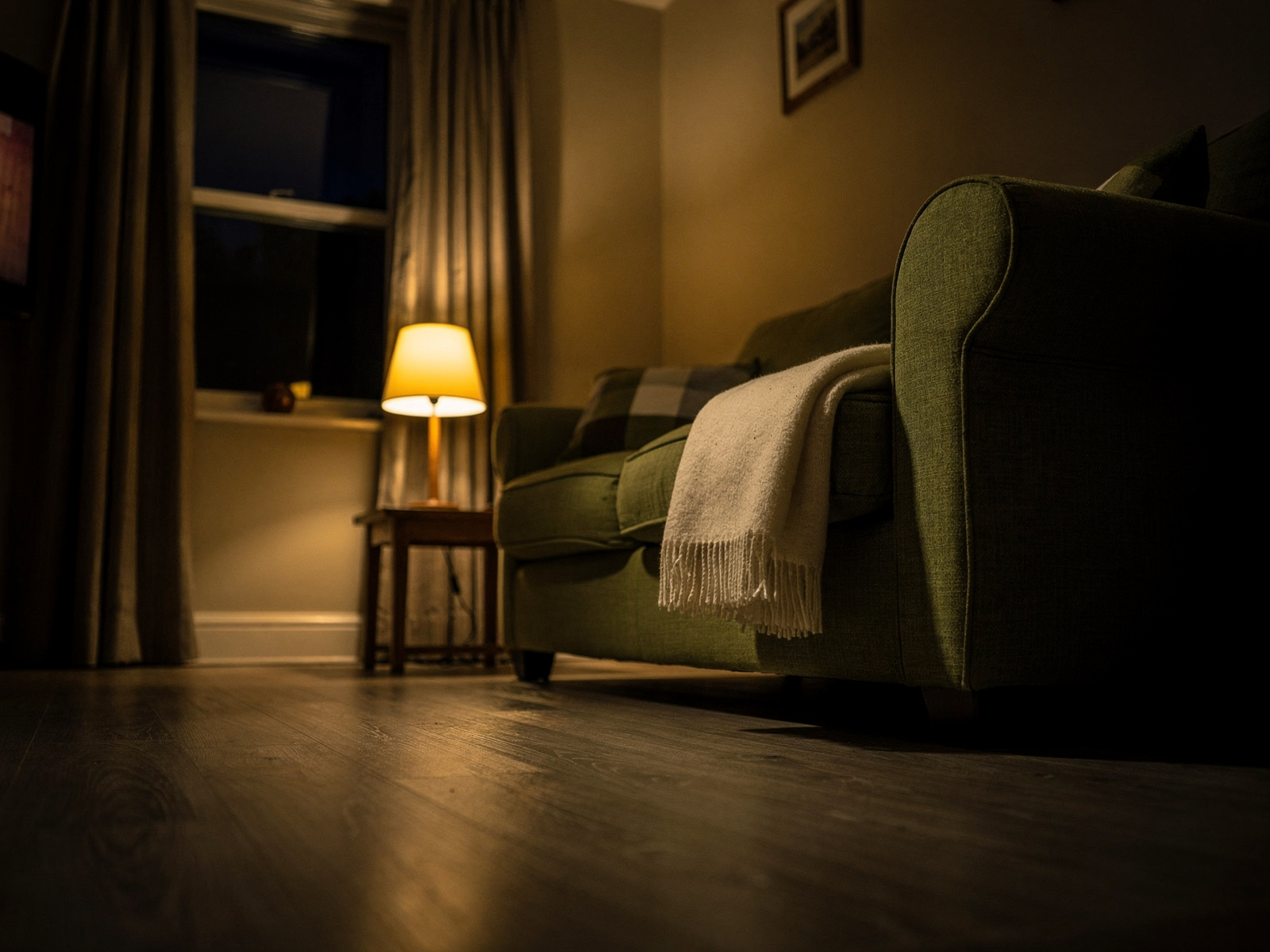 A living room in the evening with the ceiling light off and a single low side lamp glowing beside a sofa and a folded blanket.