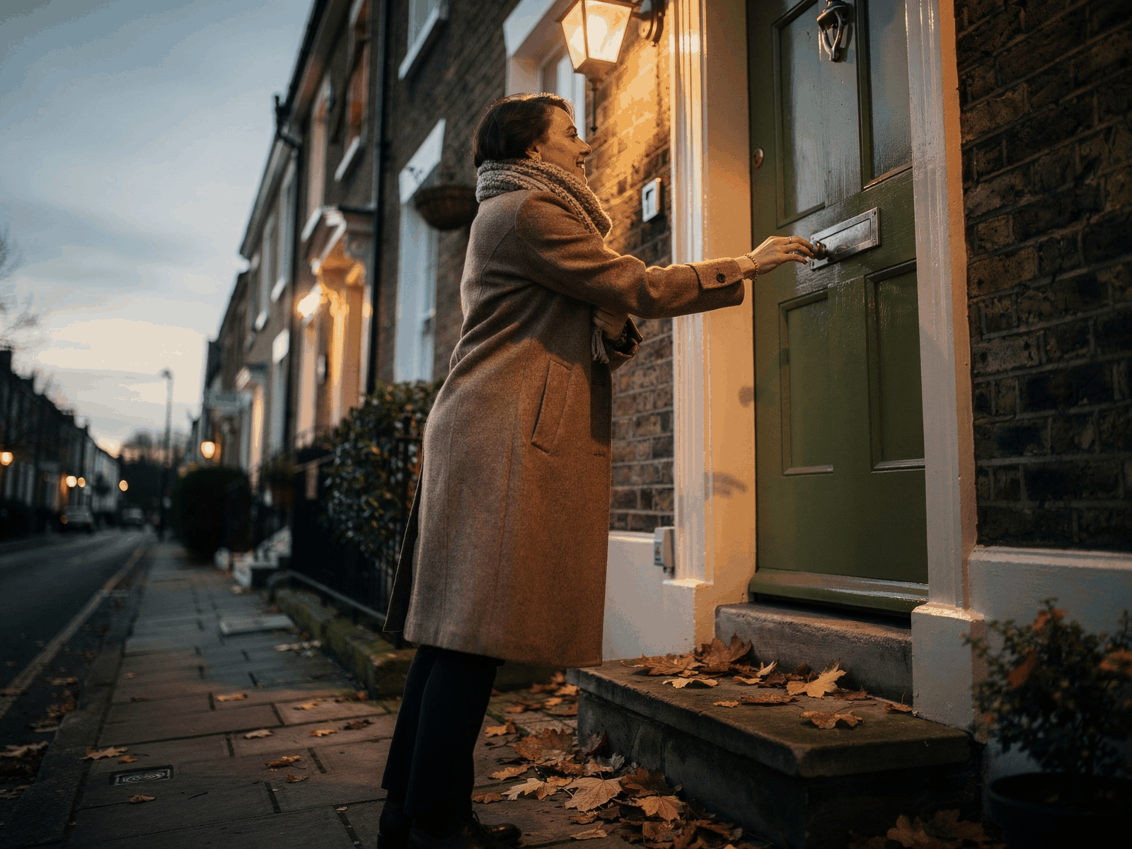 A person in a coat and scarf pulling the front door shut in late afternoon light, ready for a short walk with the street lamps just coming on.