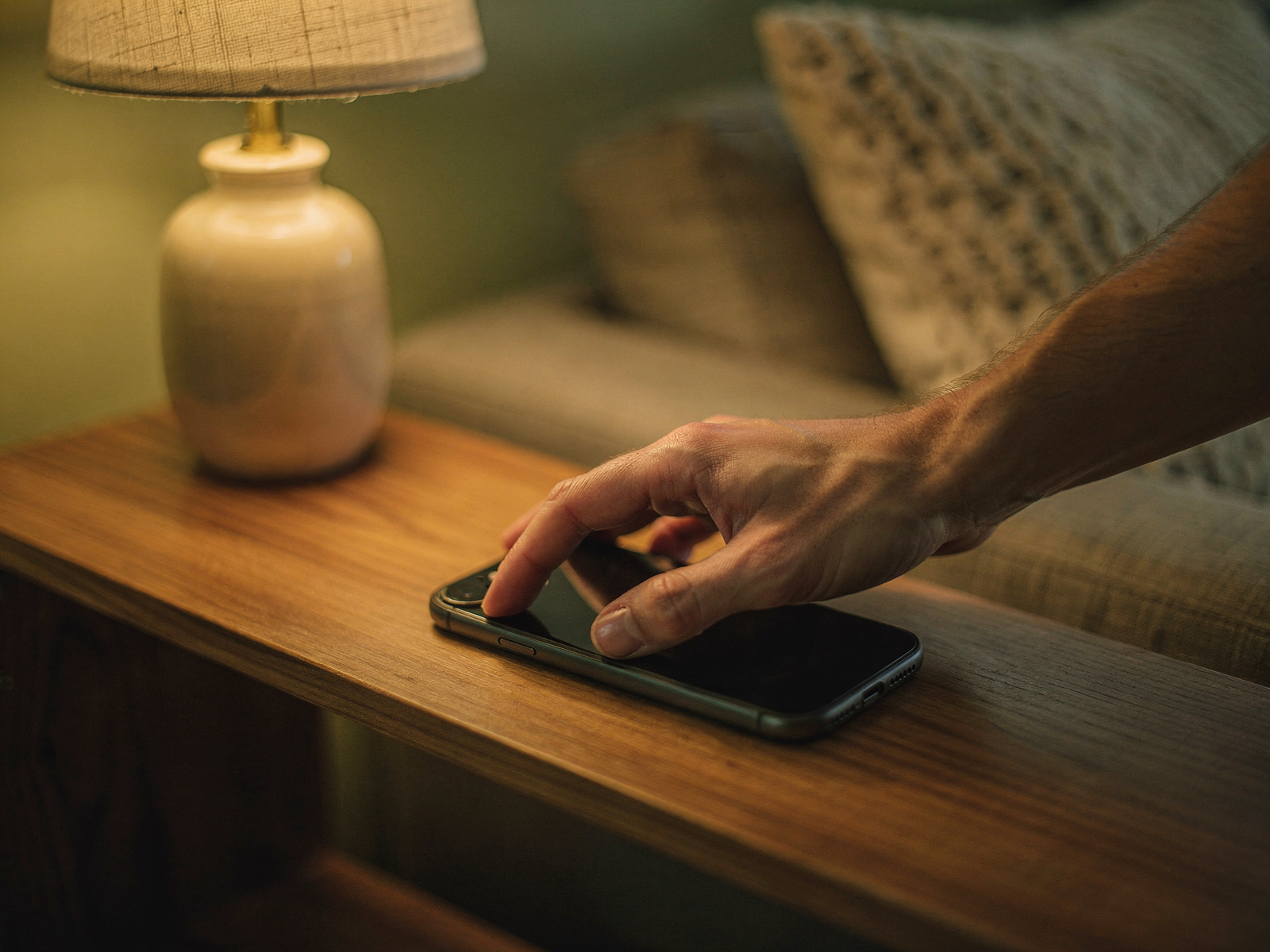 A hand placing a phone face down on a wooden shelf beside a small lamp in a softly lit living room in the evening.