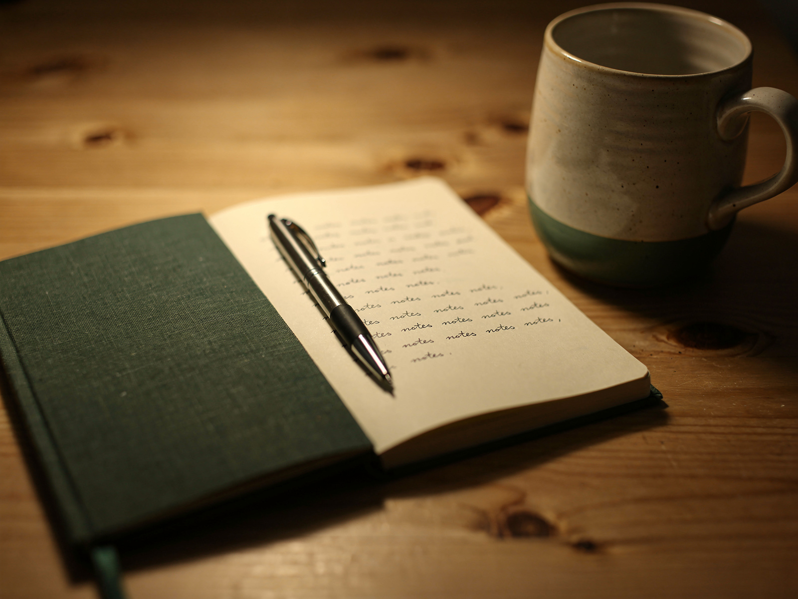 A plain notebook and pen beside a mug on a kitchen table in warm lamplight, the handwriting on the page too small to read.