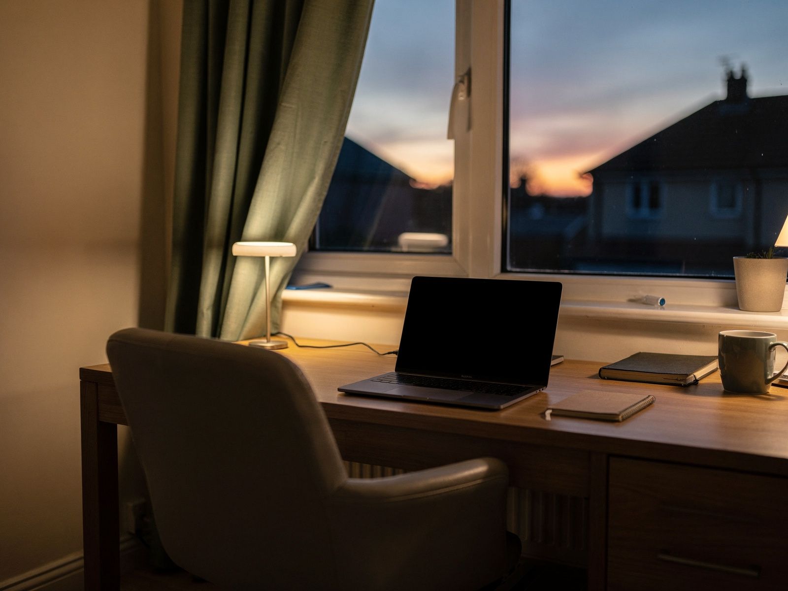 A home desk with a laptop beside a window at dusk, the room light low and a faint orange sky outside.