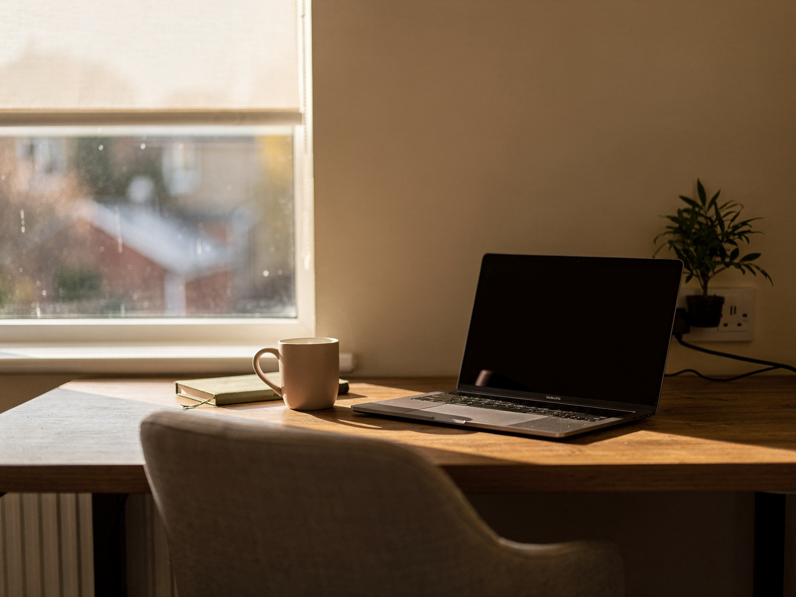 A tidy home desk set against a window with a laptop angled slightly away from the glass, soft late-afternoon light across the surface.