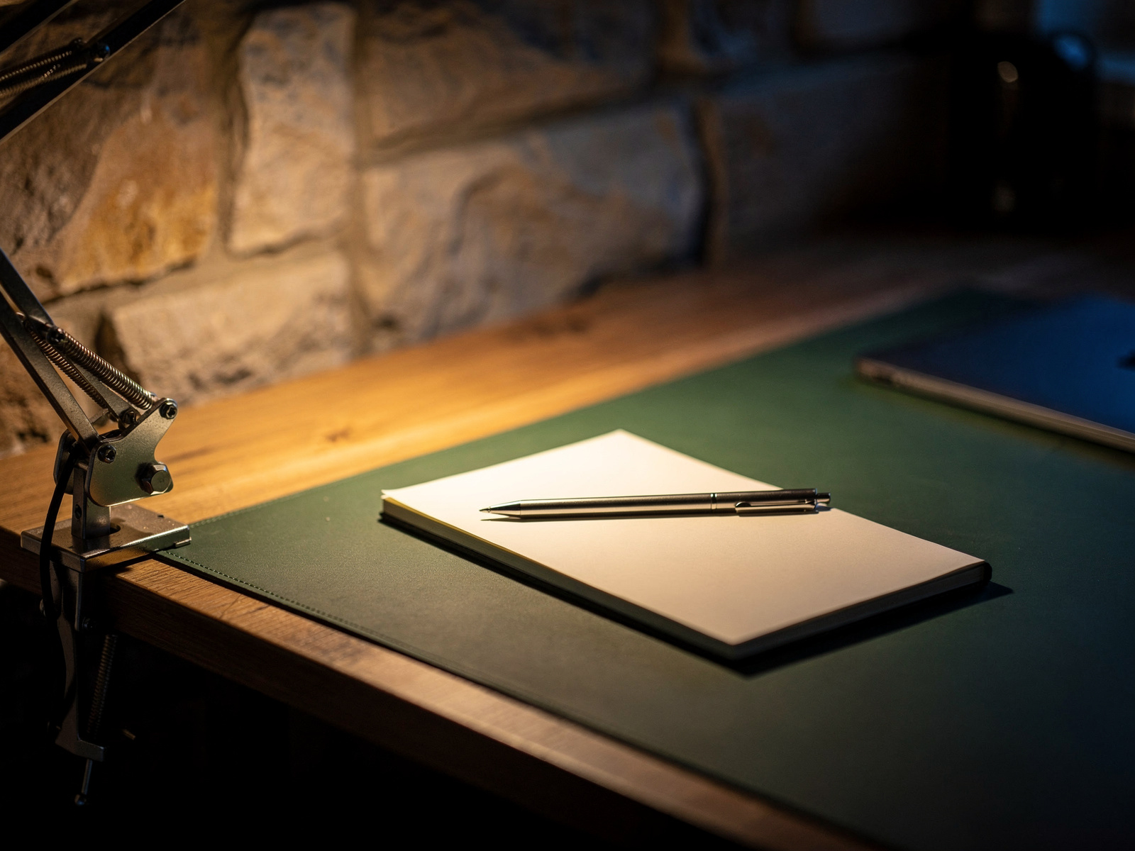 An adjustable desk lamp clamped to the edge of a wooden desk, lighting an open notebook beside a laptop in the evening.
