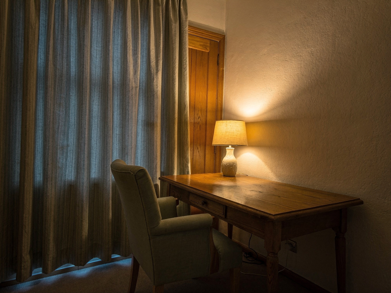 A home desk beside a window at dusk with thick curtains drawn across it and a closed door behind the chair, lit by a warm table lamp.