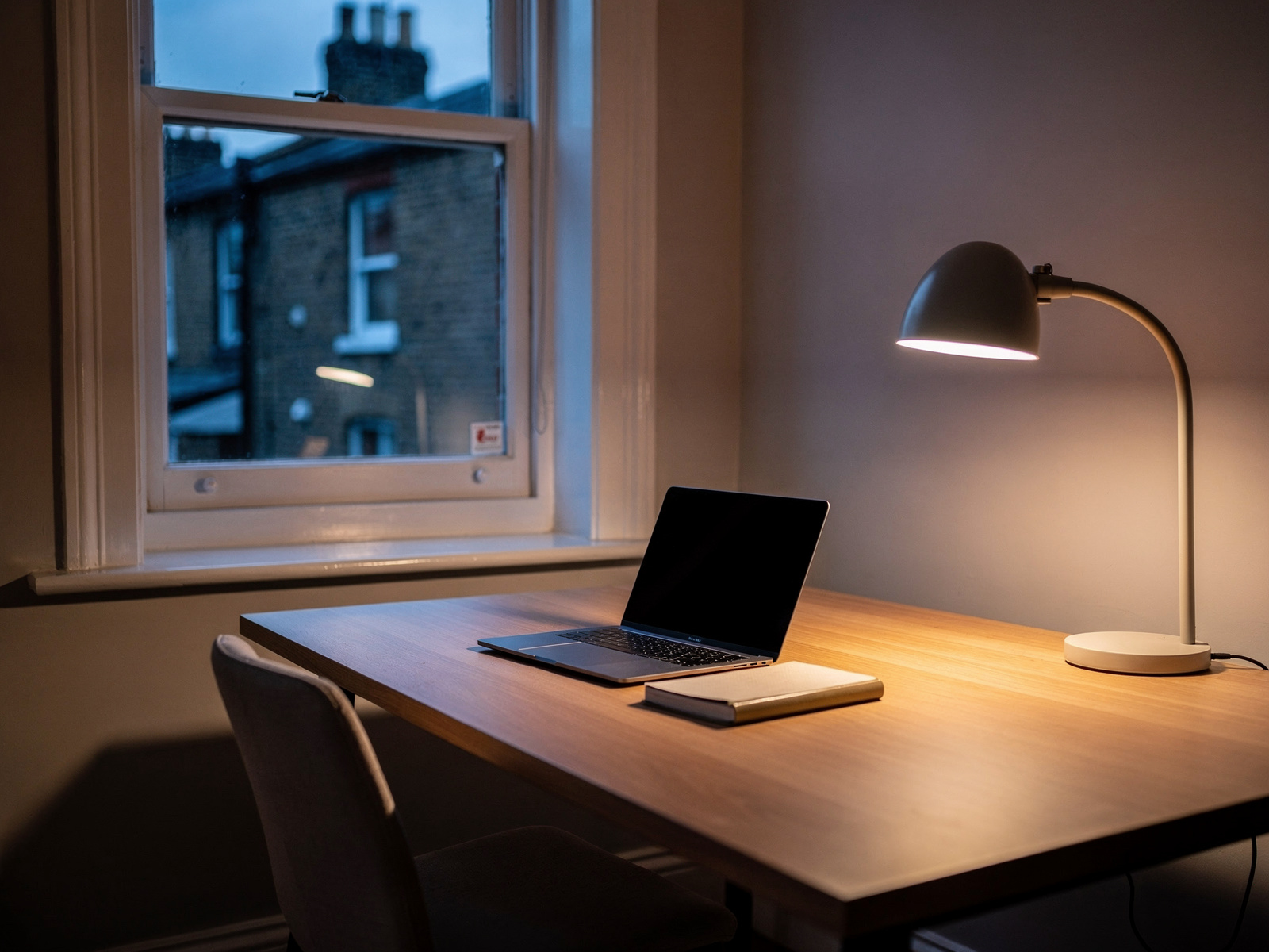 A home-working desk by a window at dusk, cleared of clutter apart from a laptop and notebook, with a gap left at the corner for a small decoration.