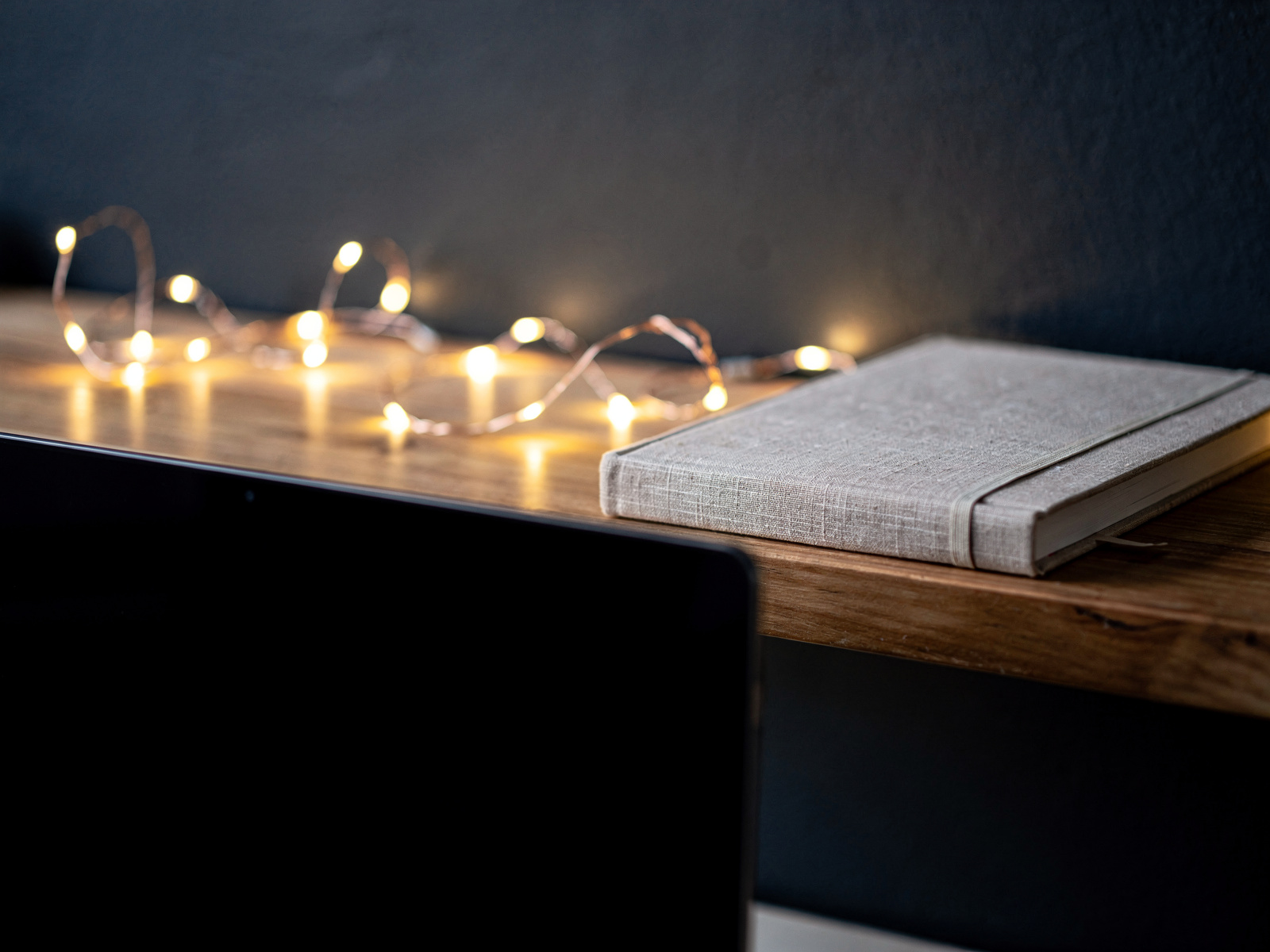 A short string of warm fairy lights along a shelf behind a home desk in the evening, glowing softly beside a closed notebook.