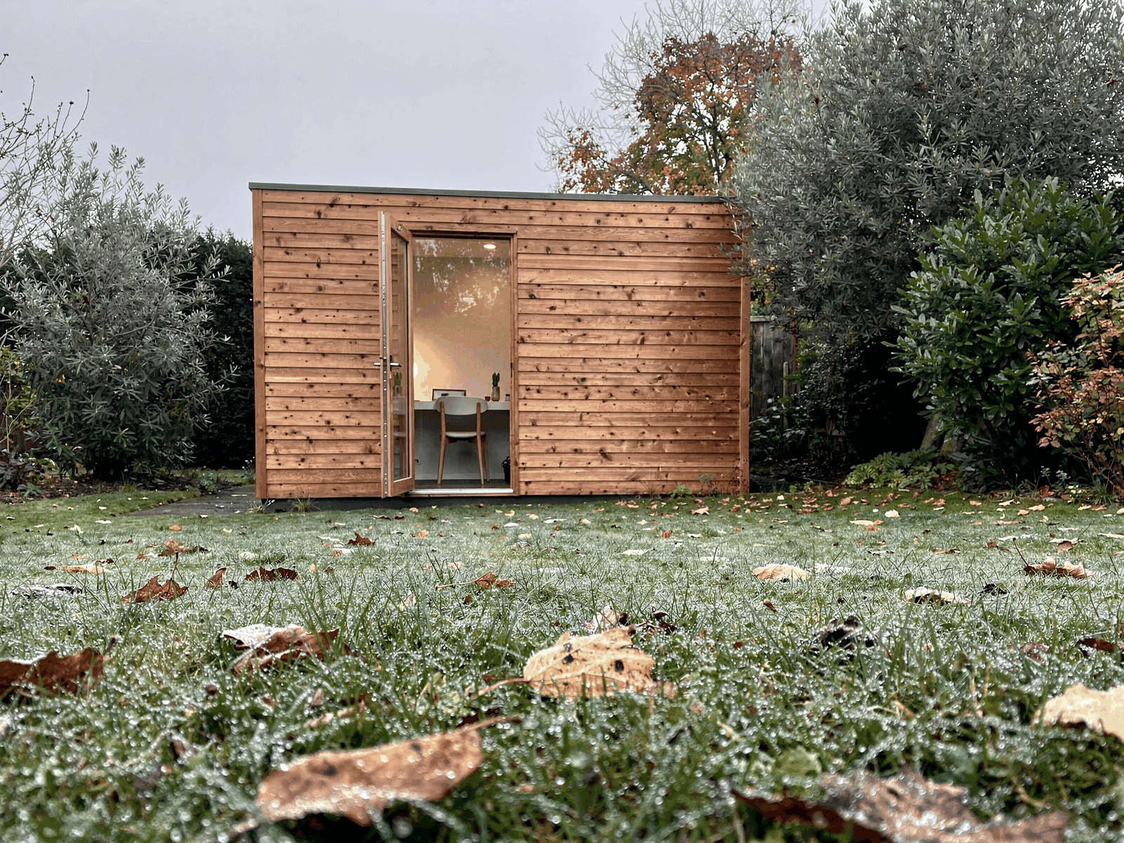 A timber garden office at the end of a lawn on a damp autumn morning, with its door slightly open and a desk visible inside.