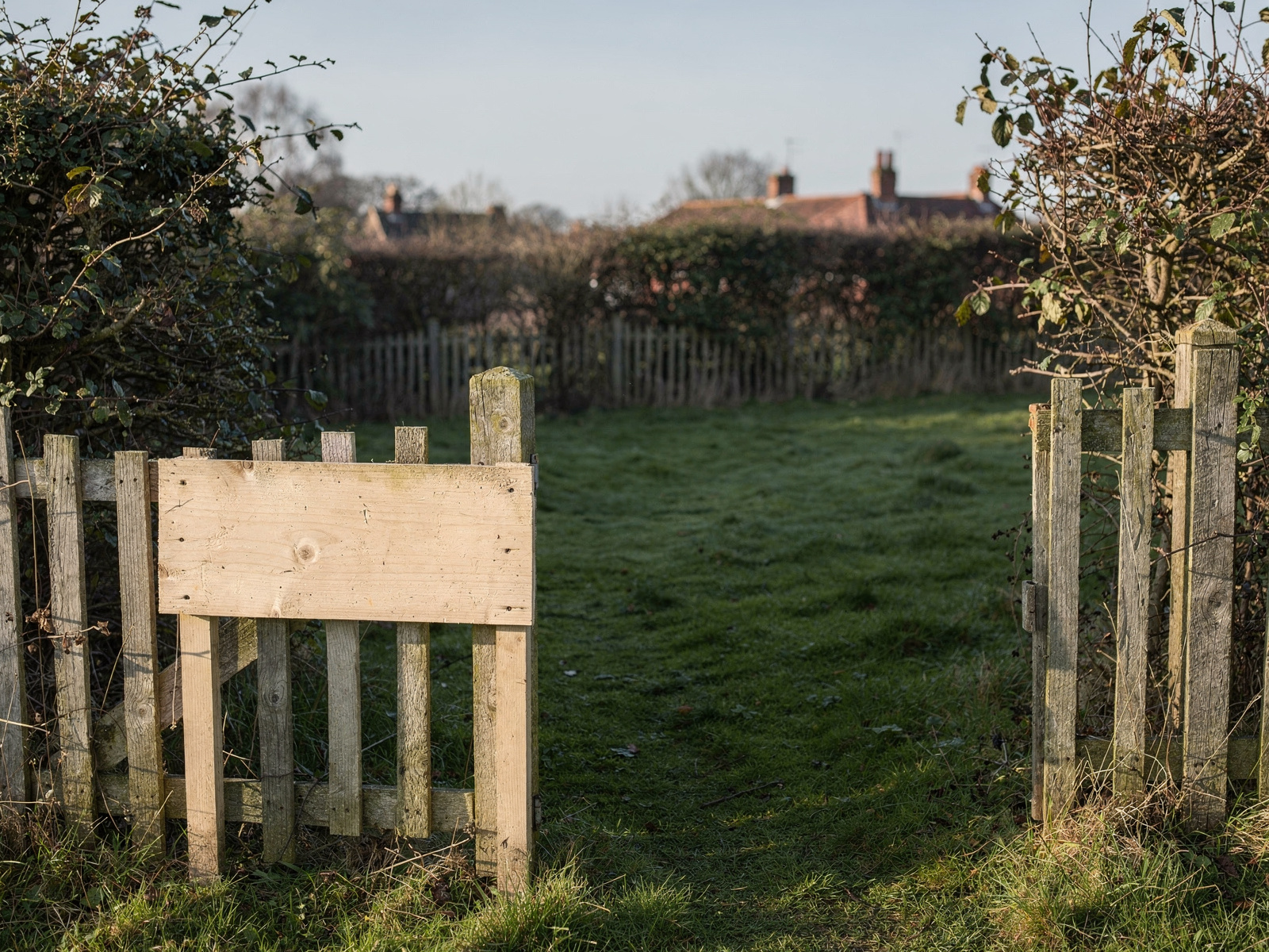 An empty fenced plot of green grass with a for-sale style signpost left blank, under a clear sky, suggesting land with nothing built on it yet.