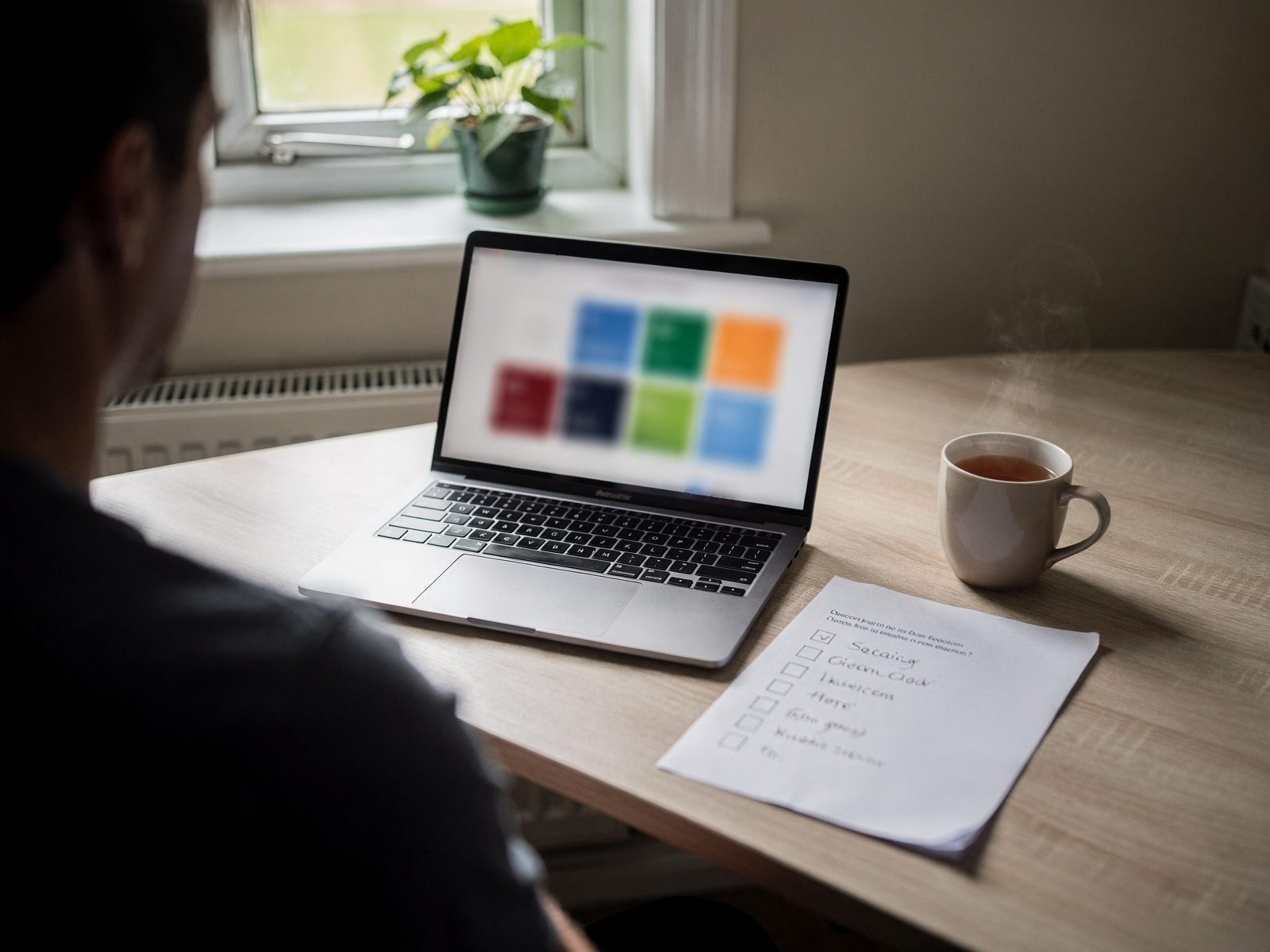 A person at a tidy home desk with a laptop open and a handwritten checklist beside a mug of tea in daylight.