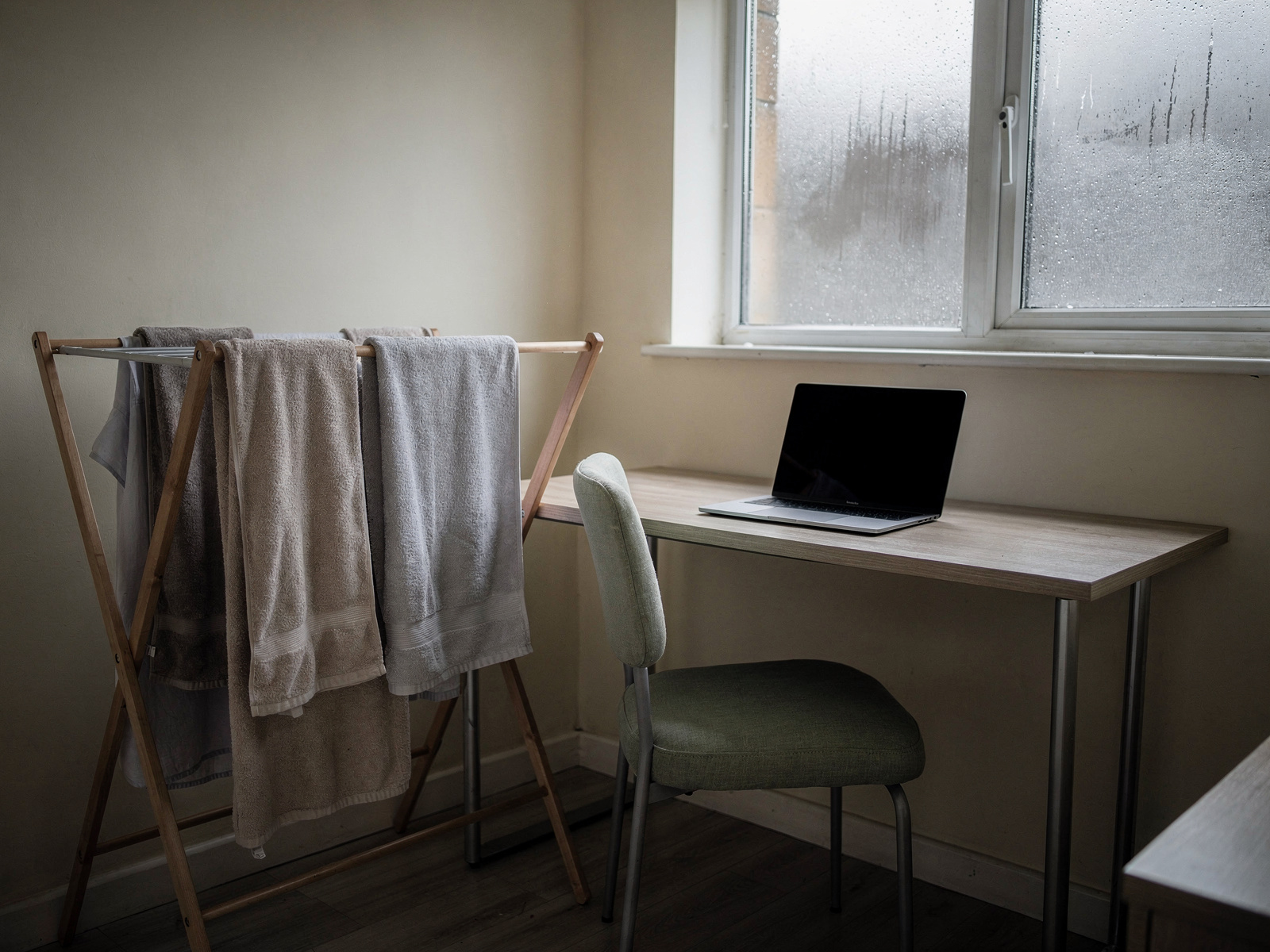 A small home office corner with a clothes airer of damp washing standing a metre from a desk and laptop, grey daylight through the window.