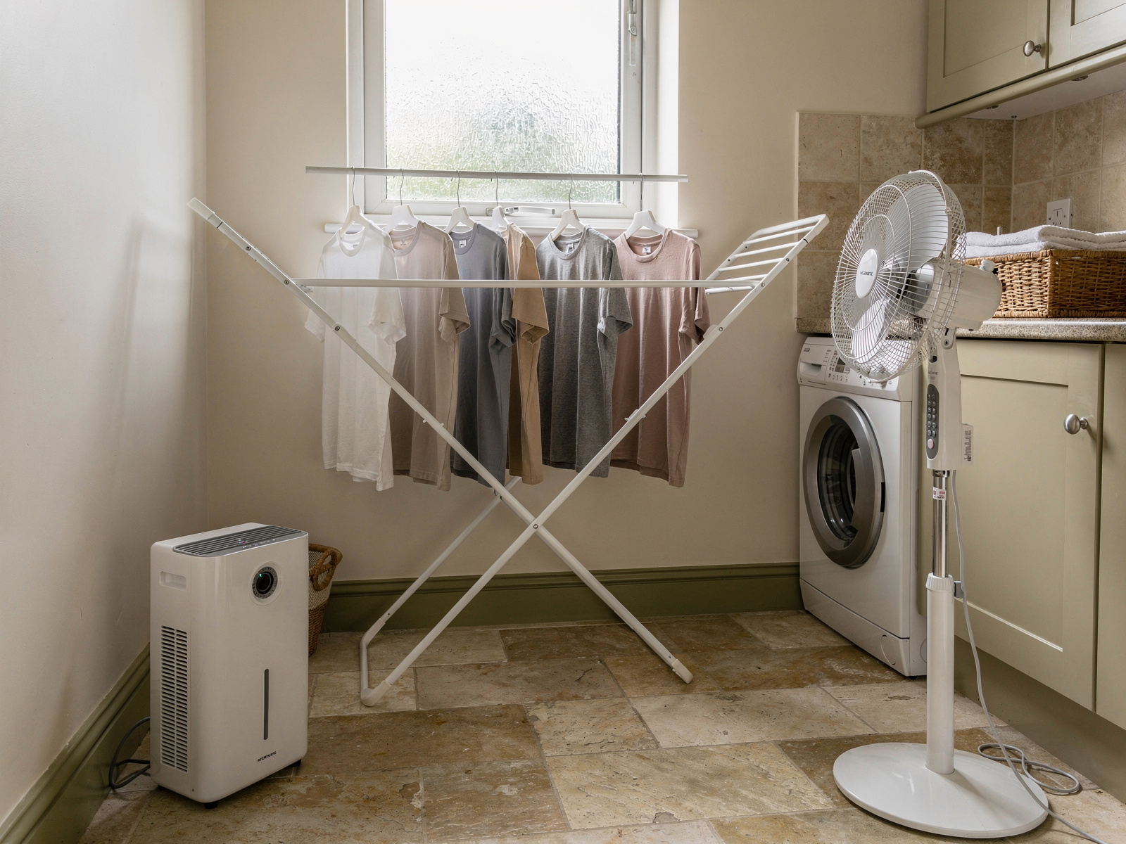 A utility room with a dehumidifier on the floor and a pedestal fan angled towards an airer of T-shirts, lit by a small frosted window.