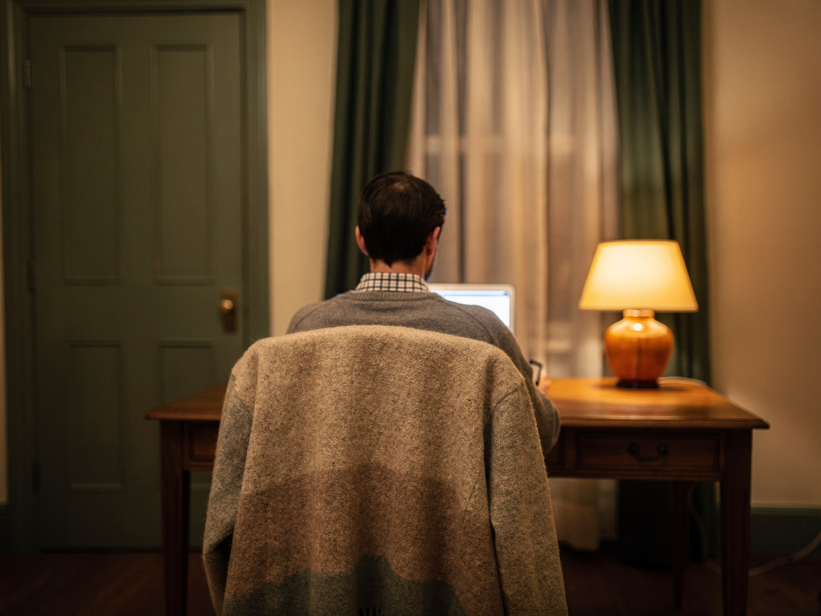 A person working at a tidy desk in a warm, quiet room in the evening, lamp on, door closed and curtains drawn.