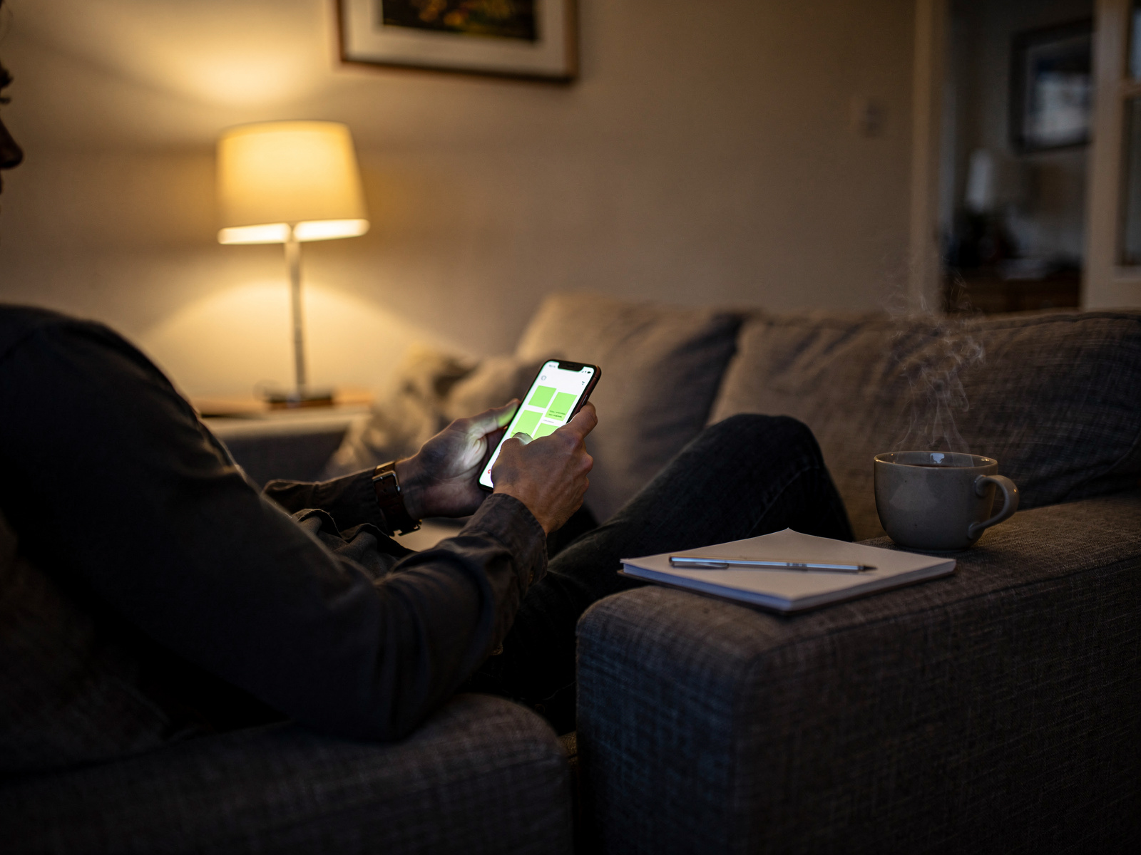 A person sitting on a sofa in the evening, reading a phone app beside a notepad with a cup of tea, warm lamp light.