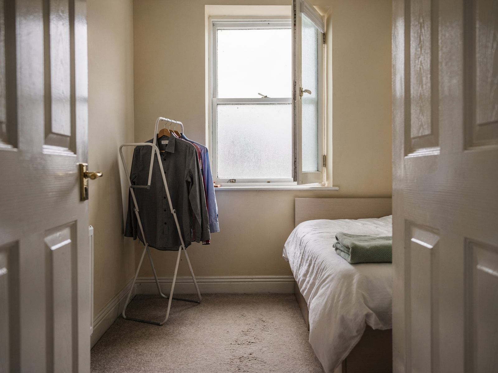 A small spare bedroom with the door closed and the window open a few inches, a clothes airer standing on the floor in soft grey morning light.