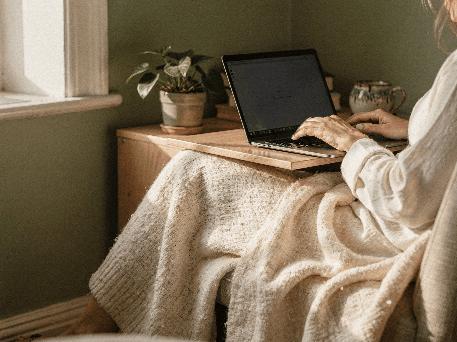 A person working at a laptop at a home desk in soft afternoon light with a throw draped over their lap.