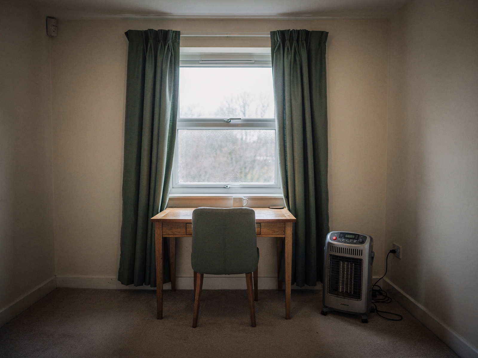 A spare-room home office on a grey afternoon, with a small desk by a window and a portable heater on the floor well clear of the curtains.