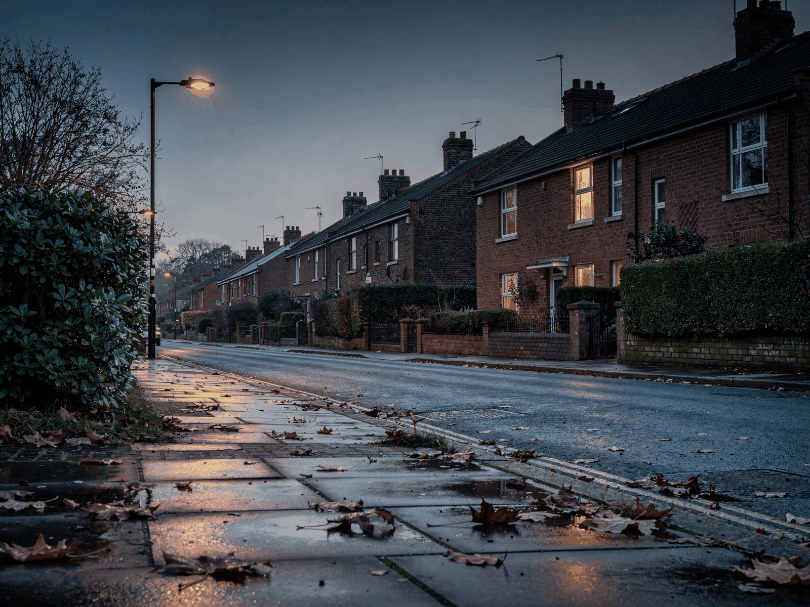 A quiet suburban street at dusk with streetlights just coming on and a lit window in the background, seen from the pavement.