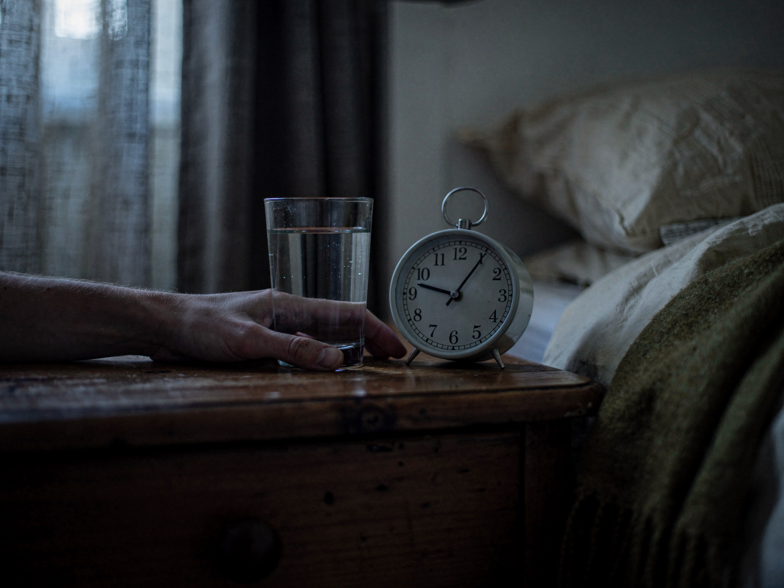 A hand resting on a bedside table next to a glass of water and an alarm clock in dim early-morning light.