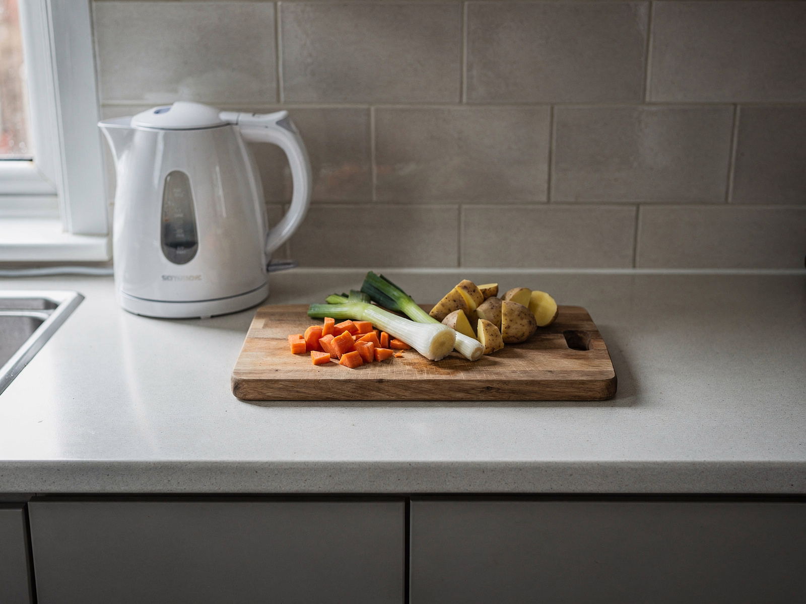 A compact kitchen worktop with a kettle, a chopping board of vegetables and a free patch of counter space, lit by a side window.
