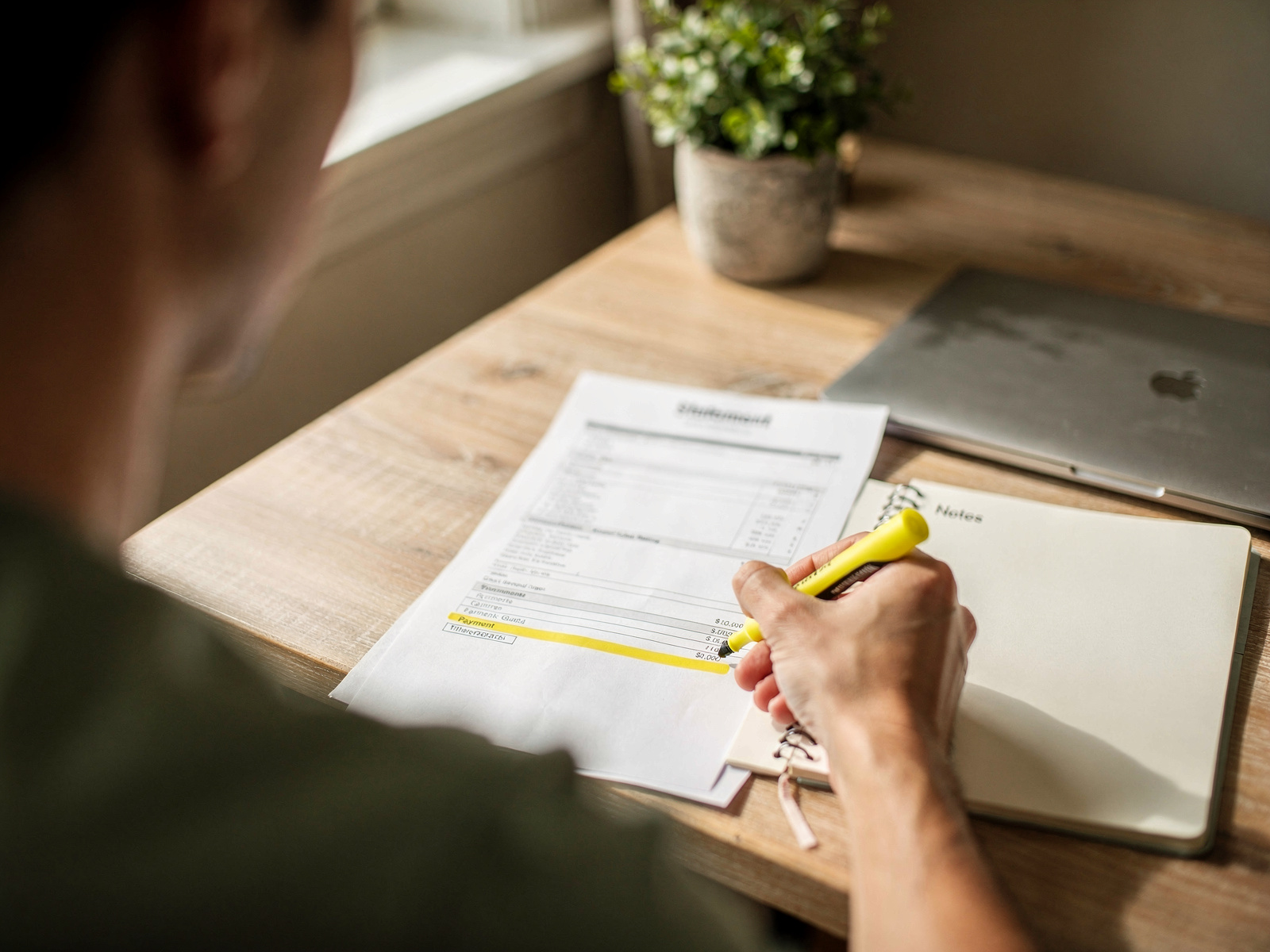 A person at a desk with a laptop, a notepad and highlighted printed statements, checking repeat payments in warm daylight.