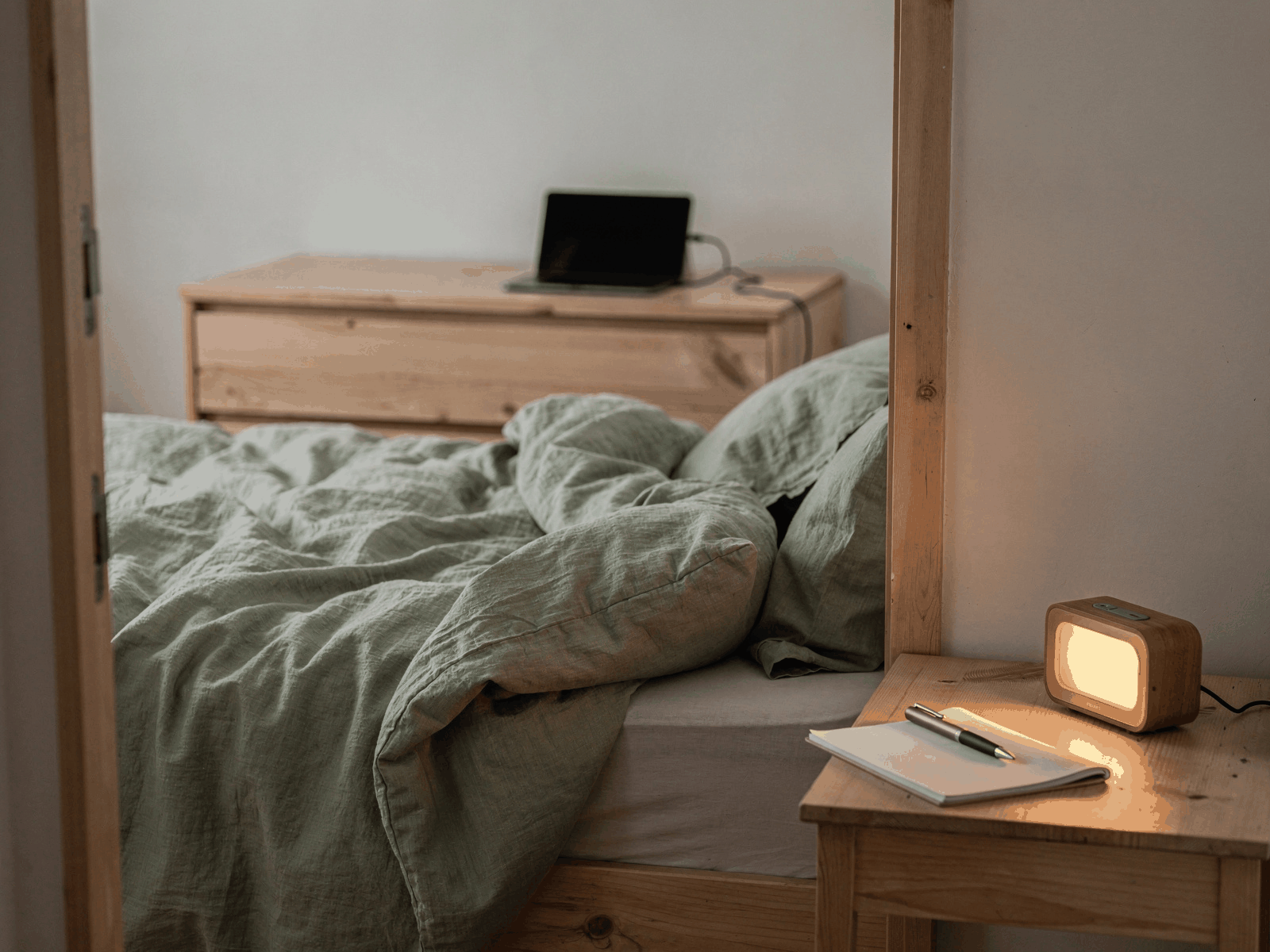 A phone charging on a dresser across a bedroom, with a small notepad and pen and a softly lit alarm clock on the bedside table.