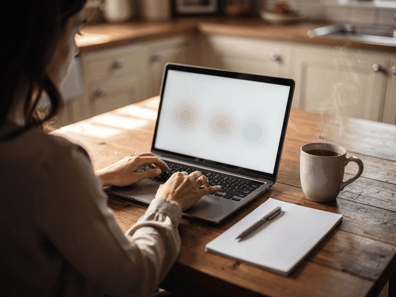 A person at a kitchen table with a notepad and a laptop open, comparing prices in daylight with a mug of tea beside them.