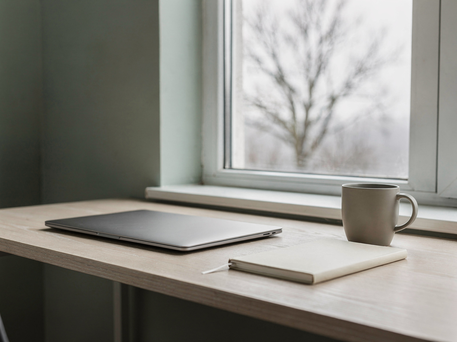A home desk beside a window in flat grey morning daylight, with a laptop, a notebook and a mug.