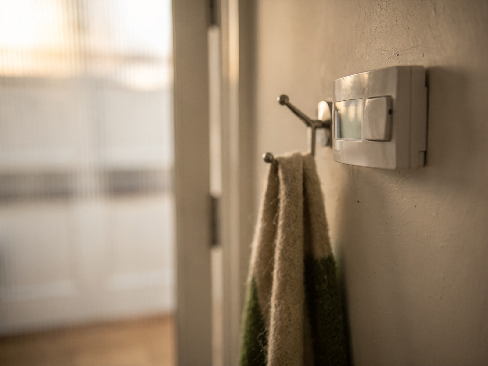 A wall thermostat in a quiet hallway in soft morning light, next to a coat hook with a scarf hanging on it.